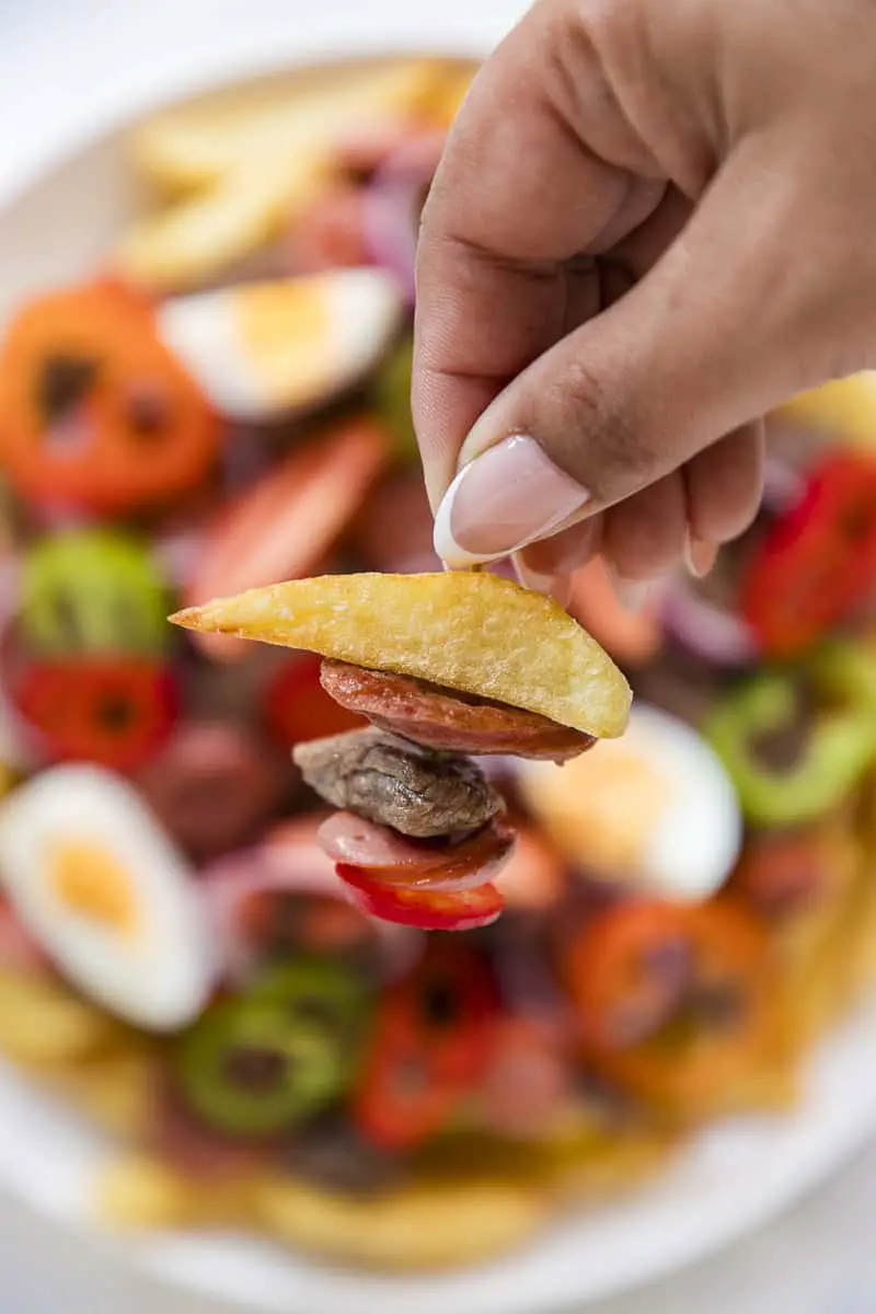 primer plano de una mano con un escarbadientes que tiene papa frita, chorizo, carne, salchicha, y aji rojo